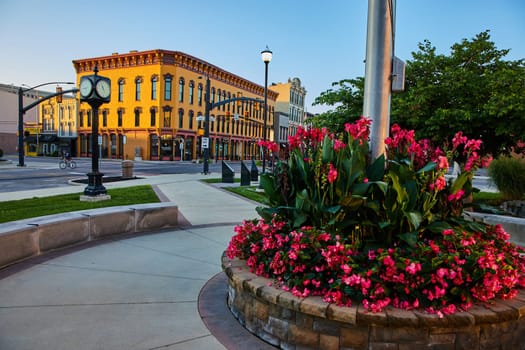 Golden hour sunrise over Muncie, Indiana in 2023, showcasing a vibrant cityscape with a blossoming flower bed, a classic street clock, and ornate buildings.