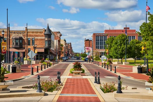 Vibrant daytime view of a charming, pedestrian-friendly main street in downtown Muncie, Indiana, 2023, showcasing a mix of traditional and modern architecture, blooming floral displays, and American flags.