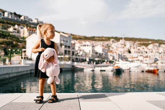 Little girl with a big pink plush hare stands on the pier against the background of the marina with yachts. High quality photo