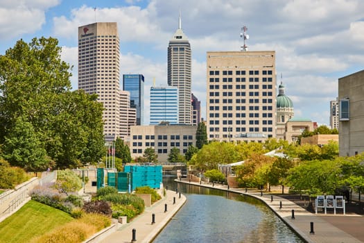 Vibrant Indianapolis cityscape with serene canal walkway amidst lush greenery, against a backdrop of modern and traditional skyscrapers, 2023