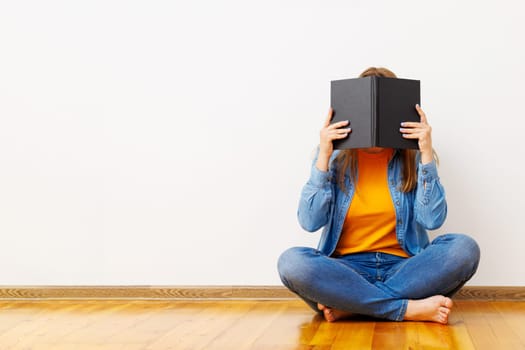 A young woman covers her face with a book as she sits on the floor against the wall.
