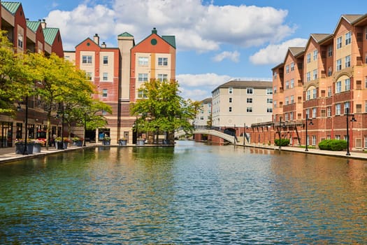 Sunny day at the serene urban canal in Indianapolis, Indiana, 2023, with modern mixed-use buildings, a pedestrian bridge, and early fall foliage.