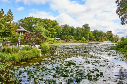 Serene lily pond with vibrant green pads, traditional gazebo, and active fountain in Botanic Gardens, Elkhart, Indiana, 2023 - a vision of tranquility and natural beauty.