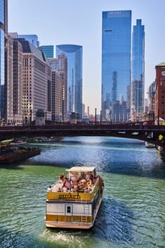 Image of Sunny tour boat, party boat on Chicago canal toward bridge on sunny day with blue skyscrapers, USA