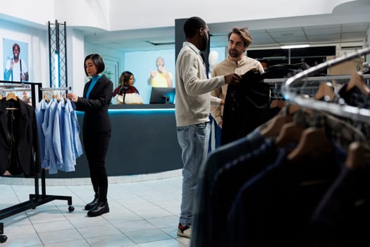 Shopping mall menswear department assistant recommending apparel size to customer. African american man consultant and caucasian buyer talking while selecting outfit in clothing store