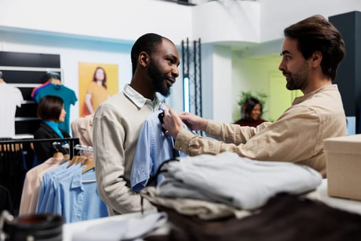 Clothing store employee and customer checking shirt and tie style match. Shopping mall boutique holding formal apparel and accessory to client body, offering fashion advice