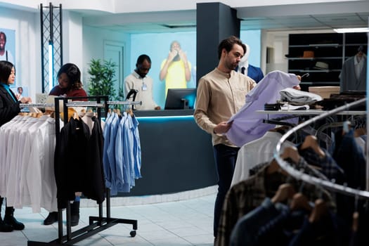 Young man holding shirt on hanger, examining fit and style while shopping in clothing store. Fashion boutique client choosing casual apparel, looking at outfit and checking size