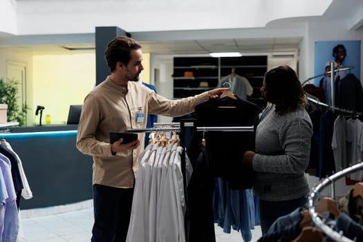 Clothing store employee showing jacket on hanger to customer, offering fashion advice. Shopping mall african american woman client and man worker choosing formal wear together