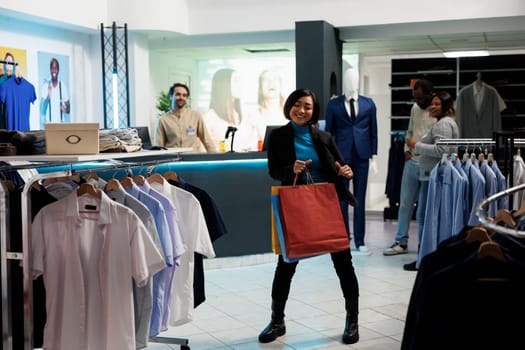 Smiling asian woman dancing with purchases, enjoying buying apparel in clothing store. Boutique cheerful customer holding packages and making moves to music in shopping center