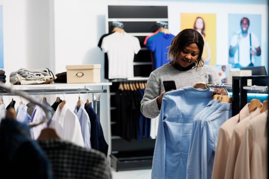 Clothing store customer choosing shirt and checking price on online website using smartphone. Smiling african american woman browsing apparel rack and scrolling shop app on mobile phone