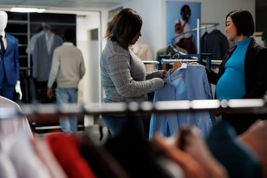 Clothing store employee and pregnant woman customer checking shirt, examining size and style. Fashion boutique smiling client and consultant chatting while choosing maternity apparel