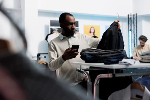 African american man choosing casual jacket in clothing store and checking price on website using mobile phone. Department mall client browsing apparel rack and examining boutique app