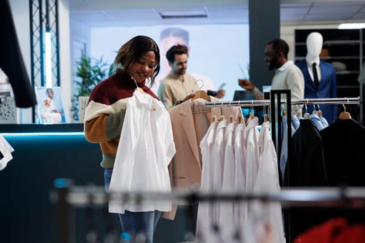 Clothing store smiling customer holding shirt on hanger and checking apparel fit. Cheerful african american woman examining trying on outfit while shopping in mall boutique