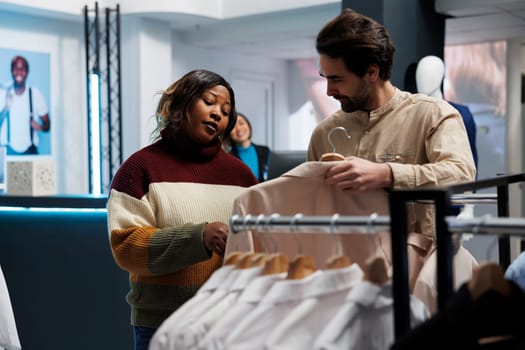 Clothing store assistant showing jacket to woman, offering fashion advice. Young caucasian man helping african american woman to choose apparel and giving recommendations in boutique