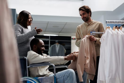 African american man in wheelchair shopping for womenswear with wife, getting help from clothing store employee. Mall customer with physical disability choosing jacket for girlfriend