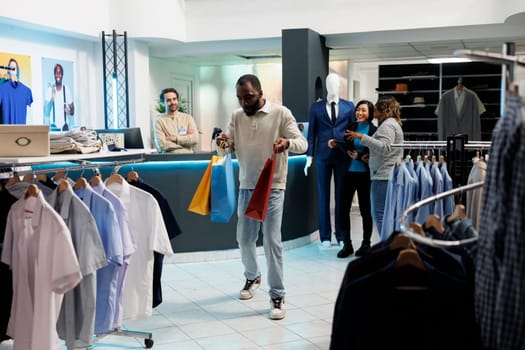 Joyful african american man carrying shopping bags and dancing after making purchase in clothing store. Fashion boutique happy customer holding paper packages filled with apparel