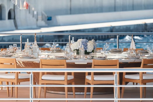 Close-up view of luxurious setting of the banquet table on an expensive mega yacht at sunny day, with awnings stretched over the deck to protect from the sun, wealth life, flowers and chairs. High quality photo