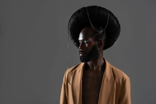 Fashion african-american bearded gay man with bright makeup and weird fashionable retro hairstyle isolated on gray background dressed beige jacket on shirtless body.