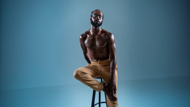 Black bearded gay wearing make-up sitting chair isolated on blue. African-american gay muscular shirtless man wearing make-up looking at camera.