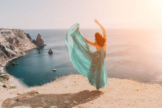 Side view a Young beautiful sensual woman in a red long dress posing on a rock high above the sea during sunrise. Girl on the nature on blue sky background. Fashion photo.