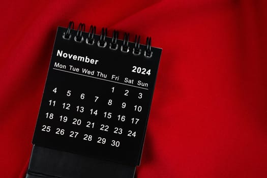 November 2024 Black spiral desktop calendar on red textile.