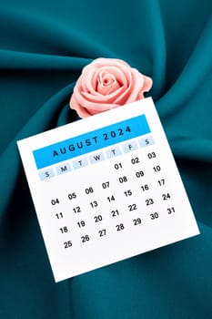 August 2024 calendar page and pink rose on green textile background.