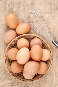 Top view of eggs in a wooden basket and egg whisk on the sack background.