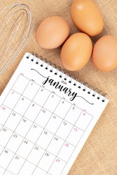 January 2024 Desk calendar and fresh eggs with egg whisk on burlap background.