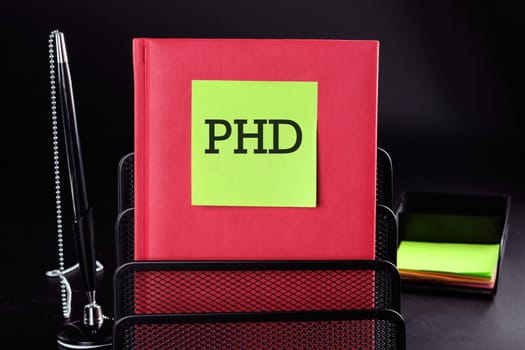PHD Text on a yellow sticker on a red vertical notepad on a black background next to a pen and money