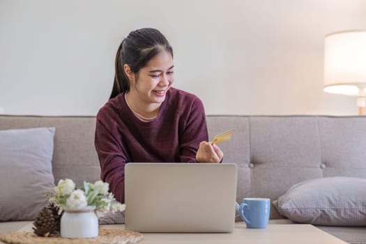 Young Asian woman in a good mood sits on the sofa in the living room and uses a laptop and a credit card. Check out the new website for online shopping..