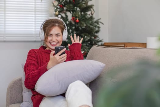 Asian woman sitting on sofa in living room using smartphone and listening to music on headphones..