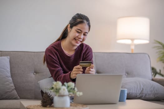 Young Asian woman in a good mood sits on the sofa in the living room and uses a laptop and a credit card. Check out the new website for online shopping..