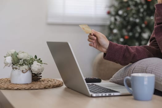 Young Asian woman in a good mood sits on the sofa in the living room and uses a laptop and a credit card. Check out the new website for online shopping..