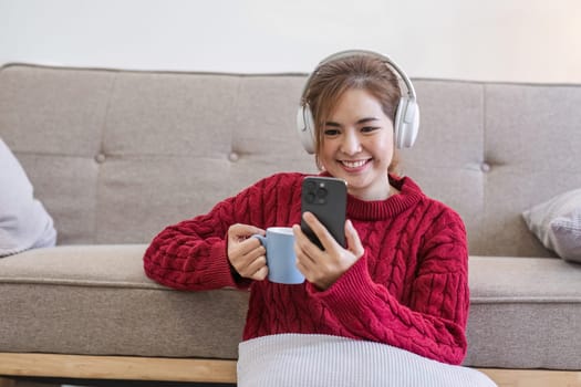 Asian woman sitting on sofa in living room using smartphone and listening to music on headphones..