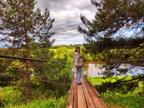 A girl on an old wooden suspension bridge among spruce or pine trees in nature. A tourist on a trip and landscape with trees on a cloudy autumn or spring day