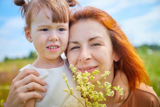 Happy female family with mother and daughter on green and yellow meadow full of grass and flower. Woman with red hair and blonde girl having fun, joy and hug in sunny summer day. Concept family love