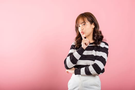 A woman in a studio shot on pink background signals quiet with a finger on her lips, inviting secrecy and mystery.