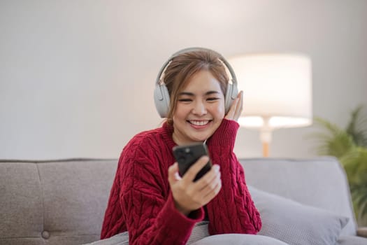 Asian woman sitting on sofa in living room using smartphone and listening to music on headphones..