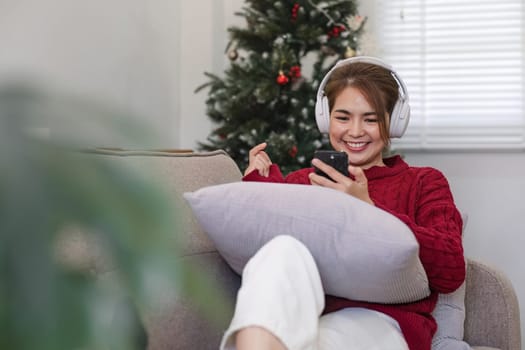 Asian woman sitting on sofa in living room using smartphone and listening to music on headphones..