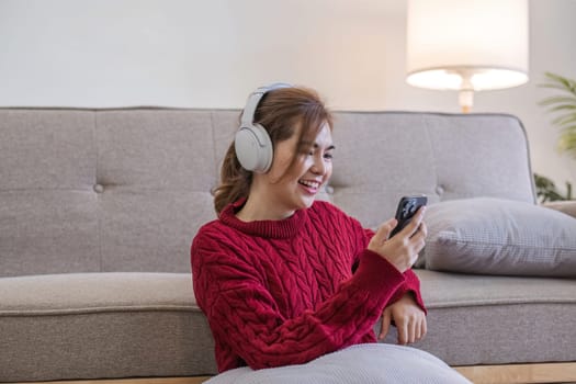 Asian woman sitting on sofa in living room using smartphone and listening to music on headphones..