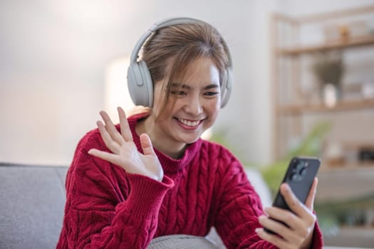 Asian woman sitting on sofa in living room using smartphone and listening to music on headphones..