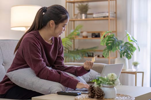 Young Asian woman in a good mood sits on the sofa in the living room and uses a laptop and a credit card. Check out the new website for online shopping..