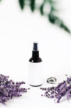 Glass spray bottle on white background surrounded by two lavender bouquets and the crystal of quartz.