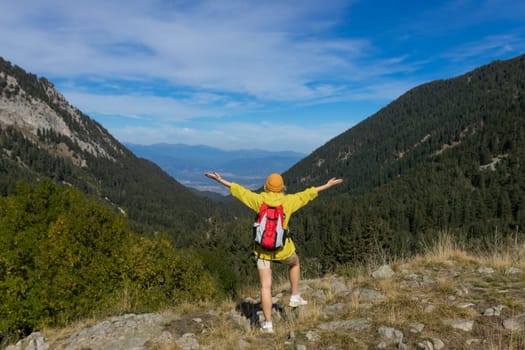 A girl on top of a mountain with green grass looks at a beautiful mountain valley. Landscape with a sporty young woman in a yellow raincoat. Travel and tourism. Hiking.