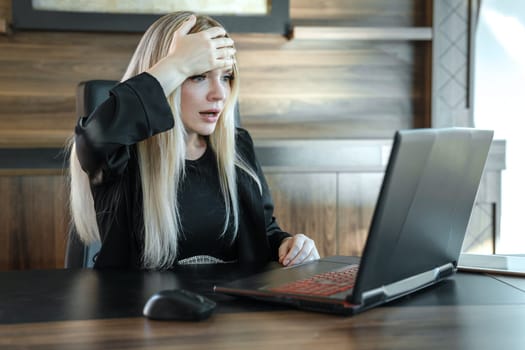 Businesswoman with frightened expression looks at laptop screen, clutching her forehead with her hand in surprise