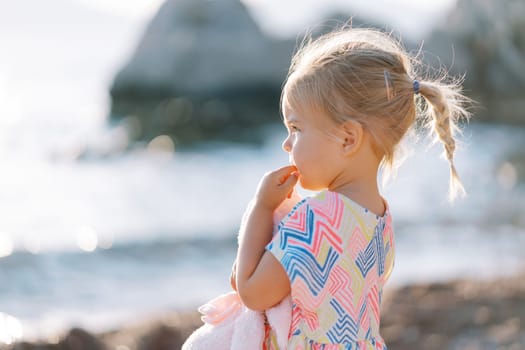 Little girl is standing on the beach with a soft toy and chewing on her finger. Side view. High quality photo