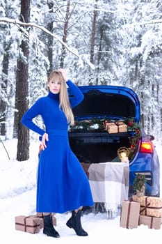 Woman in winter snowy forest in blue dress next to blue car decorated with Christmas decor. Christmas and winter holidays concept