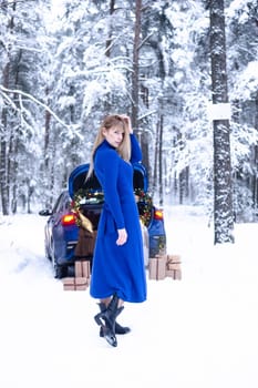 Woman in winter snowy forest in blue dress next to blue car decorated with Christmas decor. Christmas and winter holidays concept