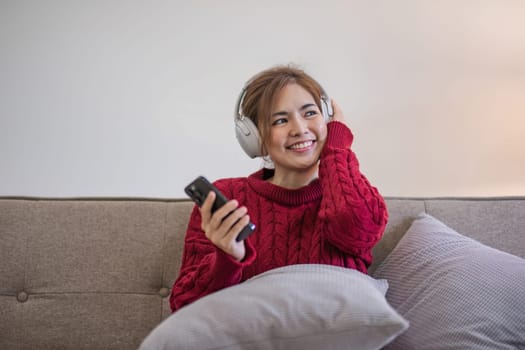 Asian woman sitting on sofa in living room using smartphone and listening to music on headphones..