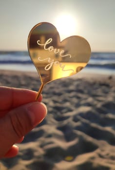 Person holding in fingers hand stick in shape heart and words Love you on background sea, waves, sky, sandy beach seashore on sunny summer day close-up. Concept love romance amour St Valentines Day